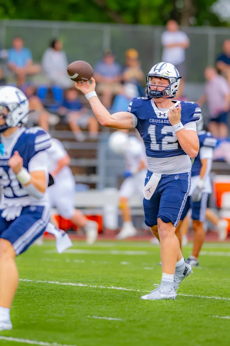 A high school quarterback in a navy jersey cocks his arm to throw a pass during a night football game